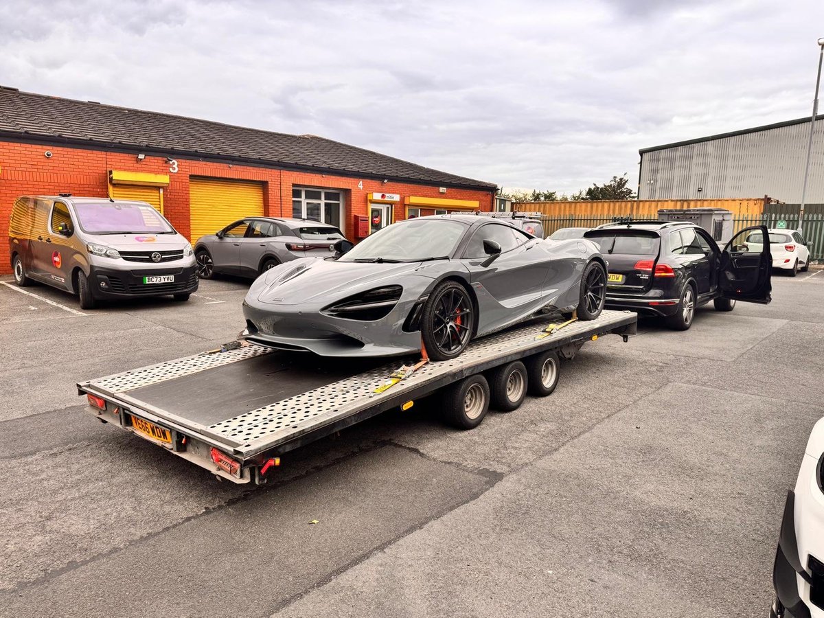 Silver sports car on flatbed trailer at auto dealership with orange building and other vehicles in background